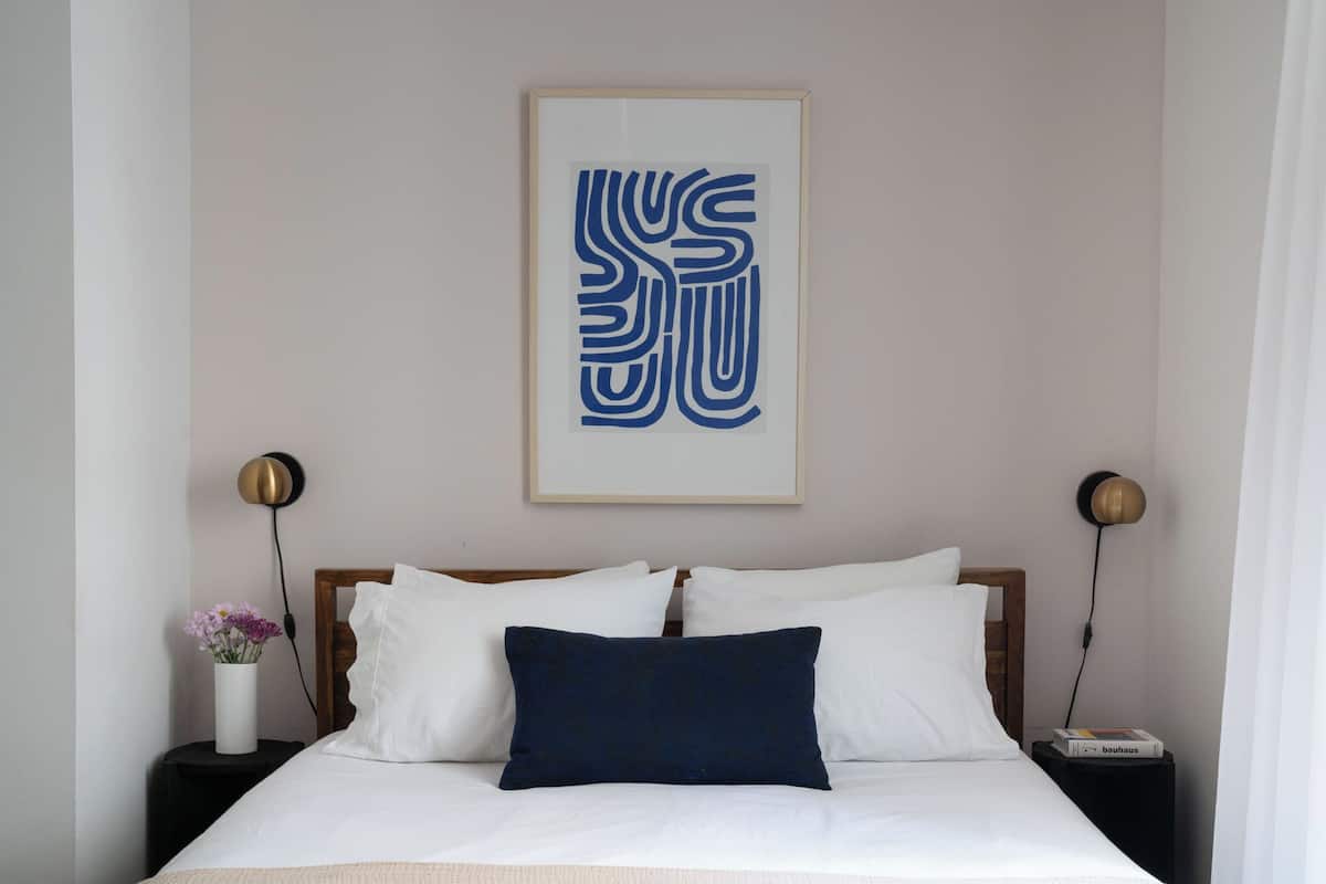 A neatly made bed with white and dark blue pillows, a framed abstract artwork above it, and two bedside lamps on either side.