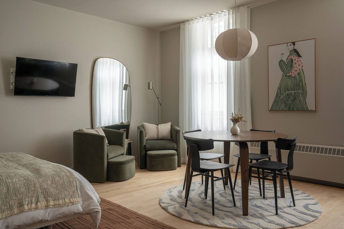 A modern room with a bed, a dining table with chairs, a sofa, a TV, and a large window with curtains.