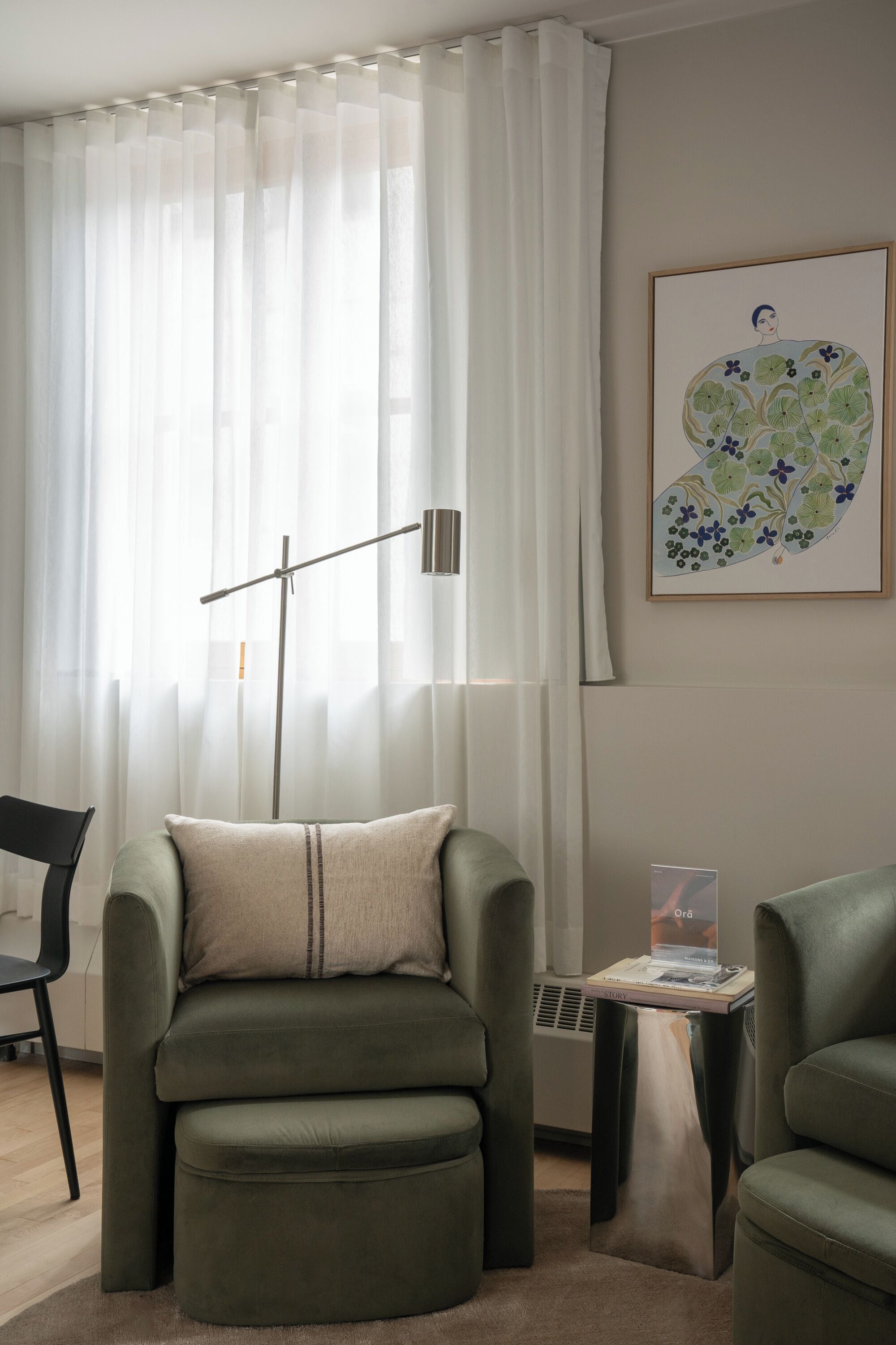 Une pièce avec un fauteuil vert, une petite table sur laquelle repose un livre, un lampadaire et une grande fenêtre ornée de rideaux légers.