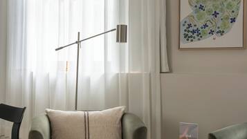 Une pièce avec un fauteuil vert, une petite table sur laquelle repose un livre, un lampadaire et une grande fenêtre ornée de rideaux légers.
