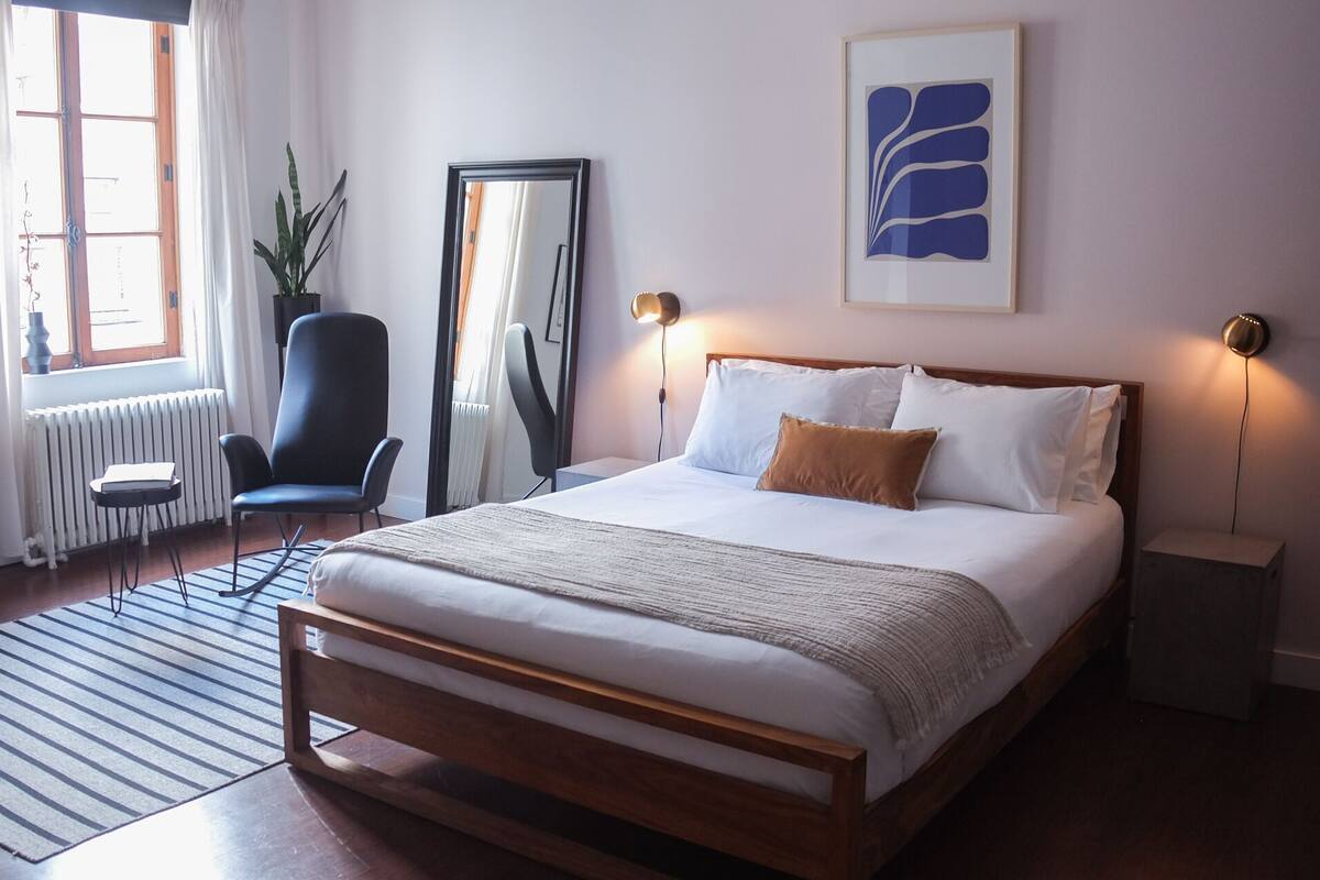 Habitación de hotel con cama de madera, un espejo grande, una planta en maceta y una obra de arte abstracta enmarcada en la pared.