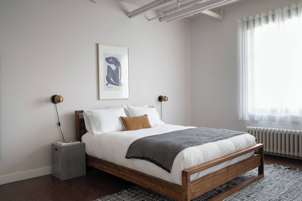 A bedroom with a wooden bed, white bedding, a gray throw blanket, and a framed artwork on the wall.
