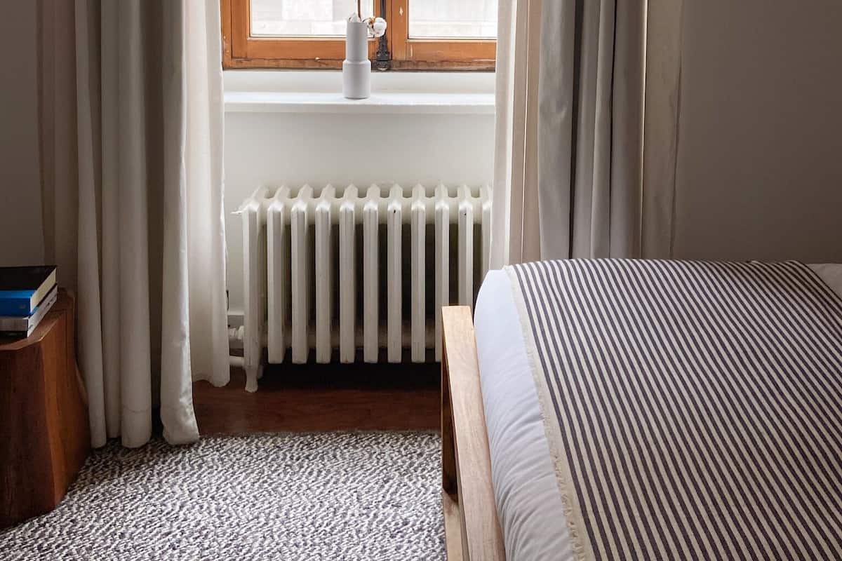 A room with a bed, a radiator, a window with curtains, and a view of buildings outside.