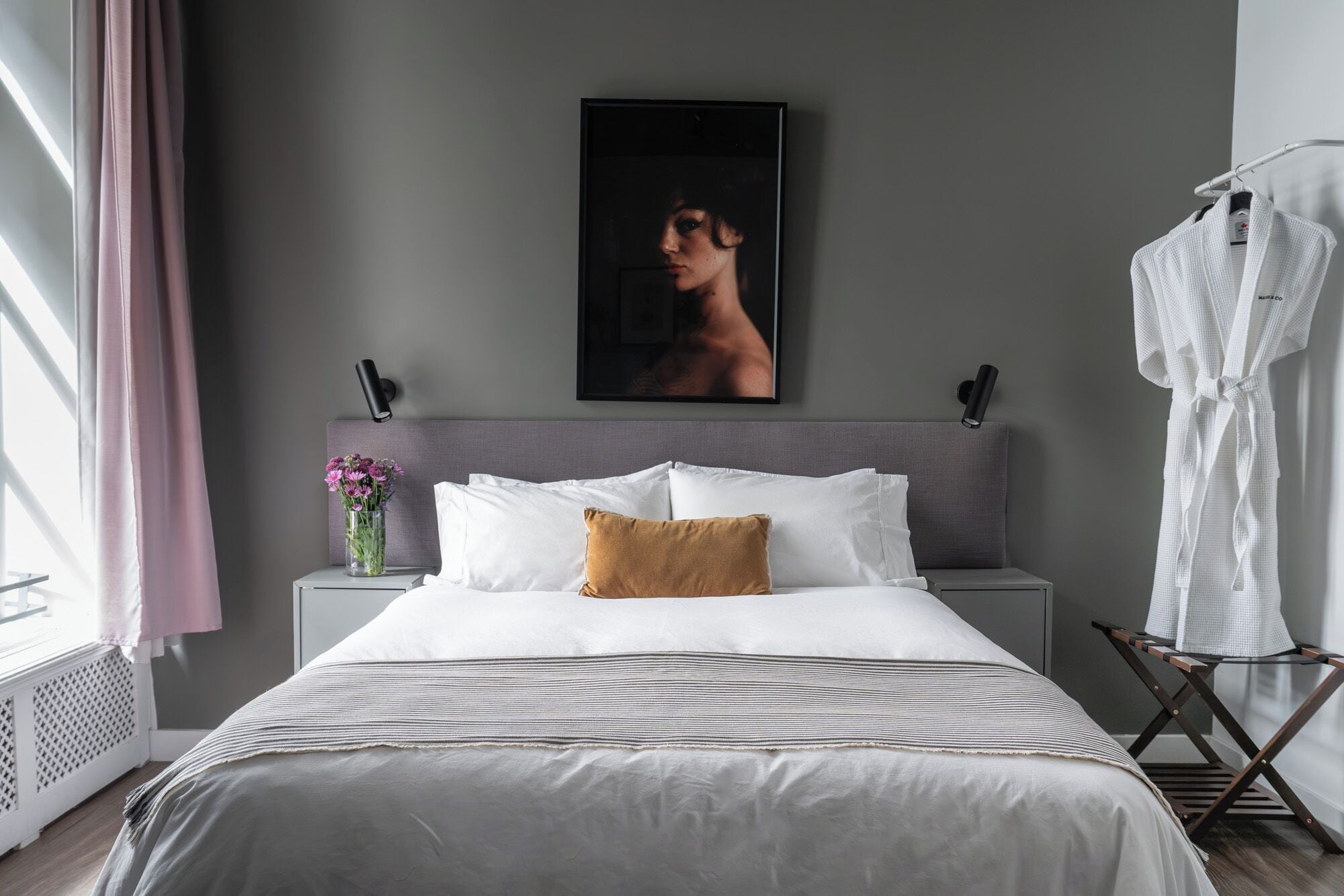 Un dormitorio moderno con una cama grande, una mesita de noche con un jarrón de flores, un cuadro en la pared y una bata colgada en un perchero.