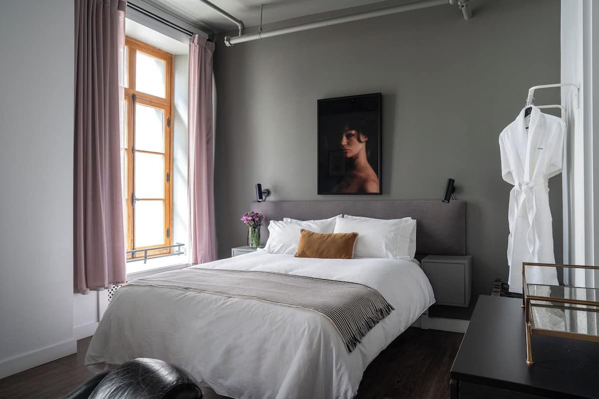 A modern hotel room with a large bed, a bedside table, a nightstand, a mirror, and a window with curtains.