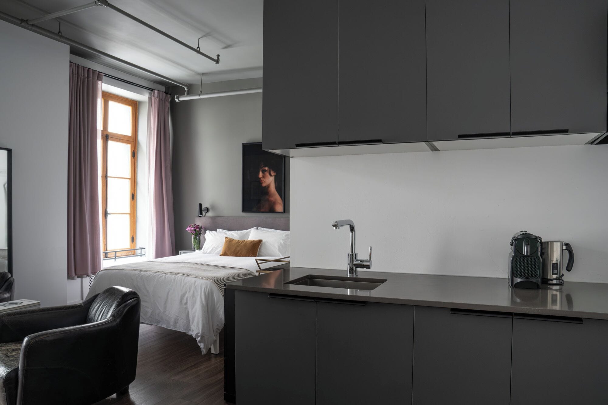 Une chambre d’hôtel moderne équipée d’un lit, d’un bureau, d’une kitchenette et d’une grande fenêtre avec des rideaux.