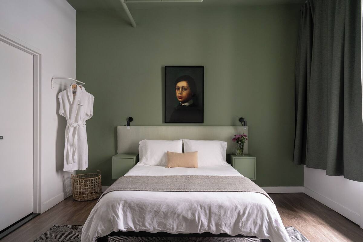 A bedroom with a bed, a nightstand, a robe, a painting, and a curtain.