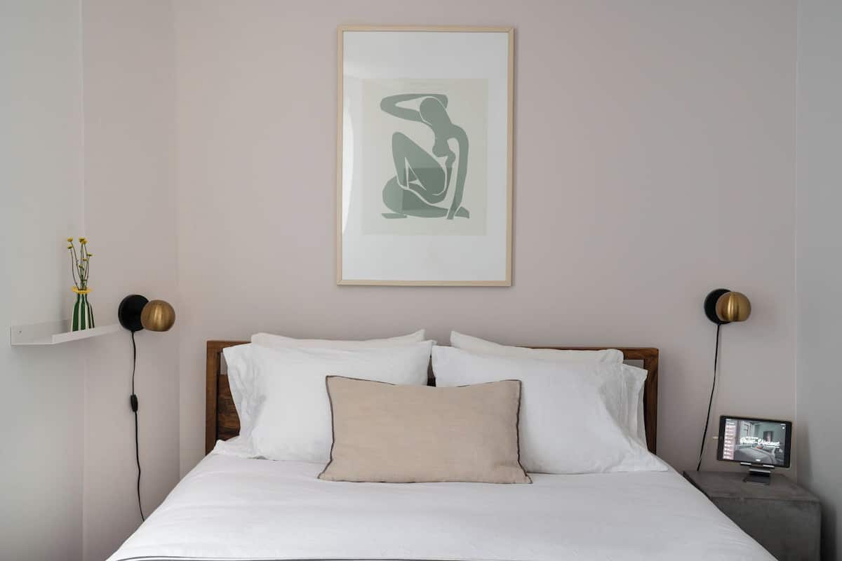 A neatly made bed with white linens and a beige pillow, a framed abstract artwork above it, and a bedside table with a lamp and a small screen displaying a car image.