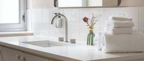 A bathroom with a sink, mirror, towel stack, toilet paper, and a vase with flowers.