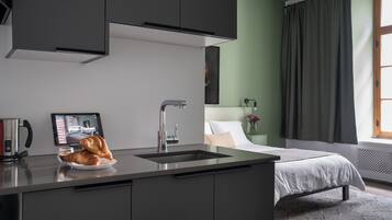 A modern hotel room with a kitchenette, a bed, a bedside table with a lamp, and a window with curtains.