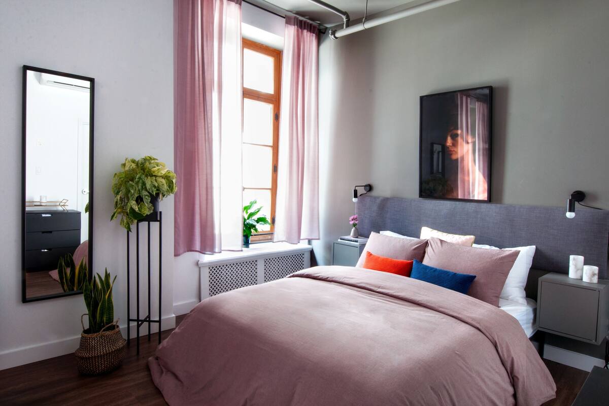 Un dormitorio con cama, mesita de noche, cortinas en la ventana, espejo y planta en maceta.