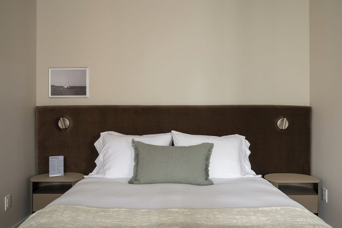 A neatly made bed with a brown headboard, white pillows, and a green accent pillow. There is a framed picture on the wall above the bed.