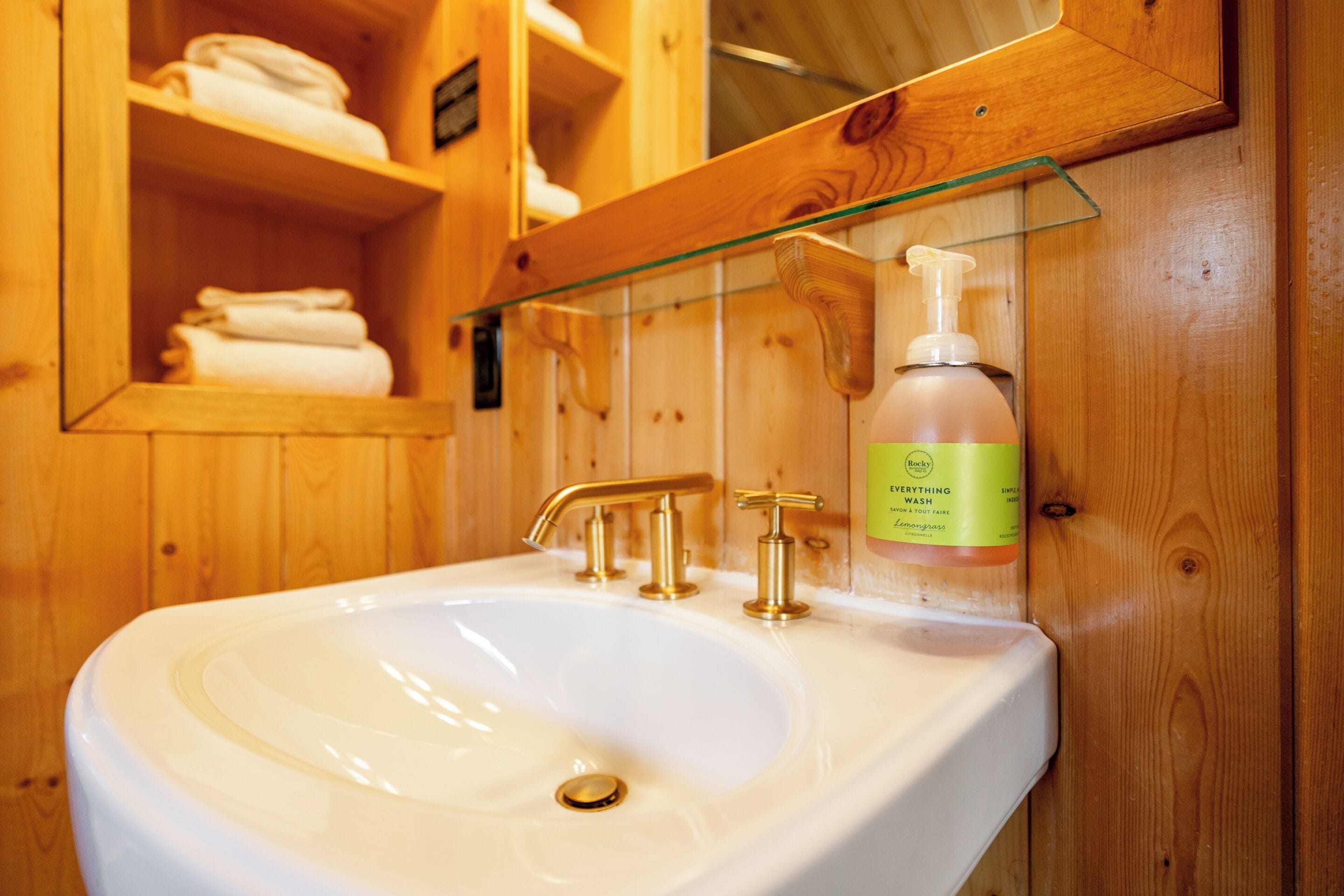 A bathroom with a white sink, gold faucets, and a soap dispenser on a wooden shelf.