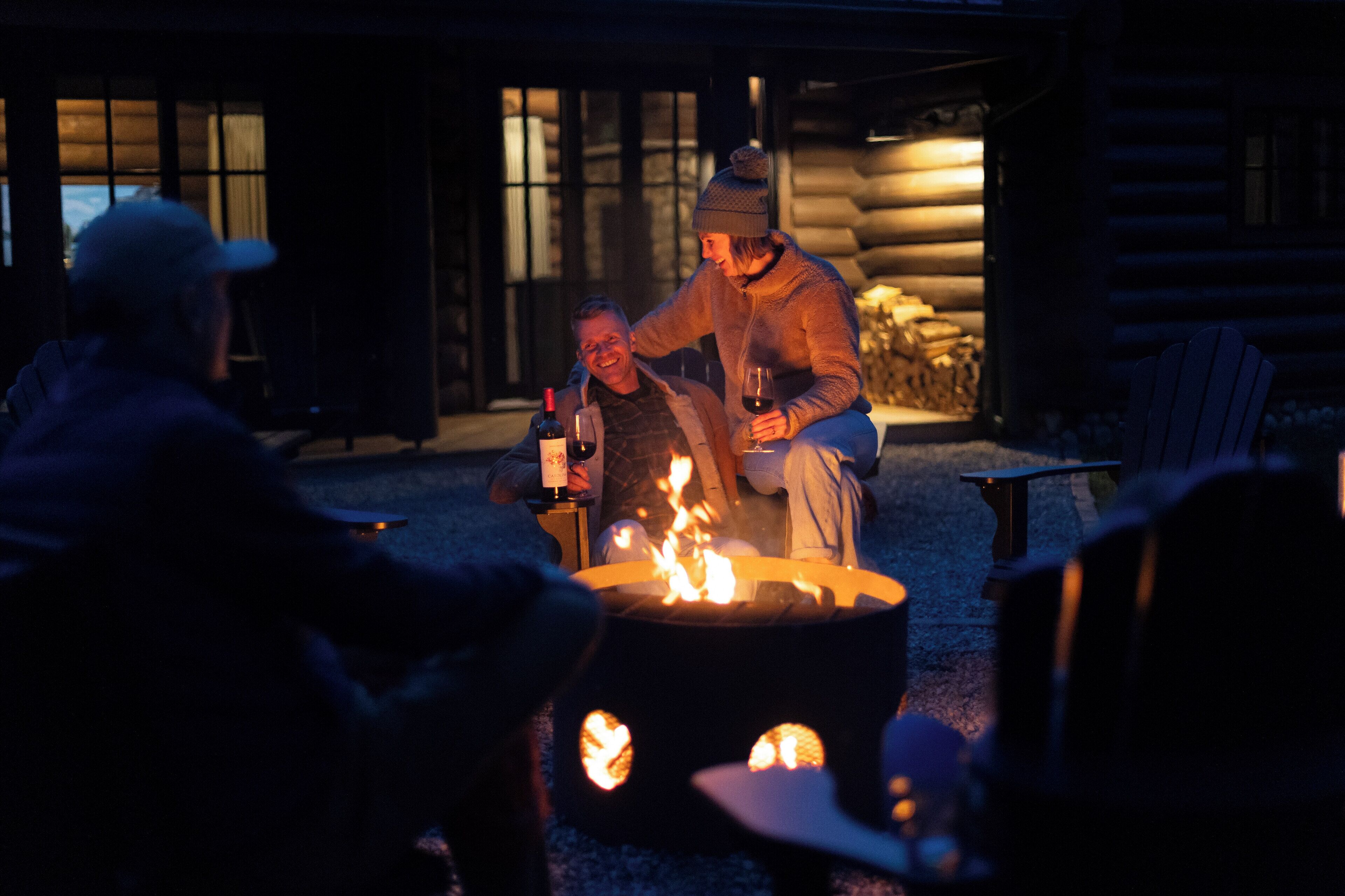 A cozy outdoor seating area with a fire pit, wooden furniture, and a rustic cabin-style building in the background.