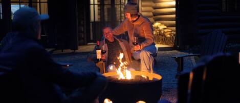 A cozy outdoor seating area with a fire pit, wooden furniture, and a rustic cabin-style building in the background.