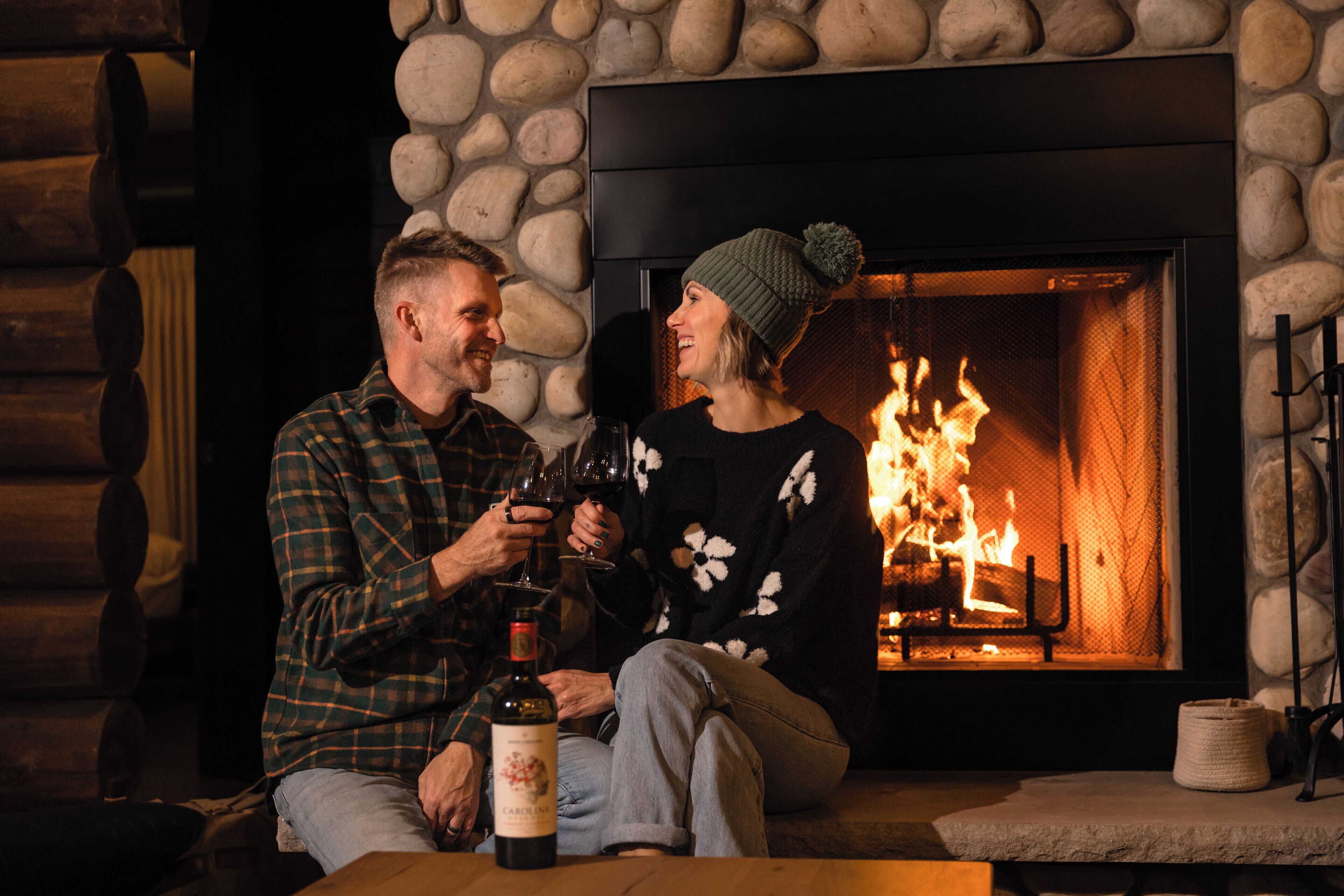 A couple sitting by a fireplace with a bottle of wine on a table.