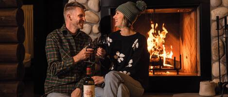 A couple sitting by a fireplace with a bottle of wine on a table.