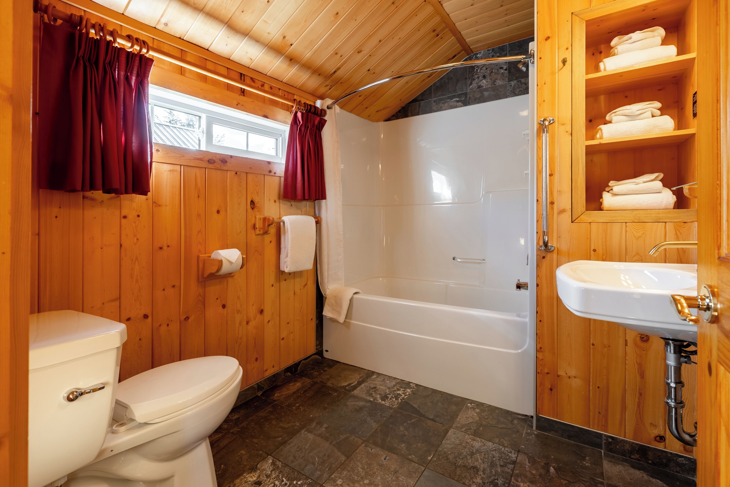 A wooden bathroom with a toilet, bathtub, and wooden shelves with towels.