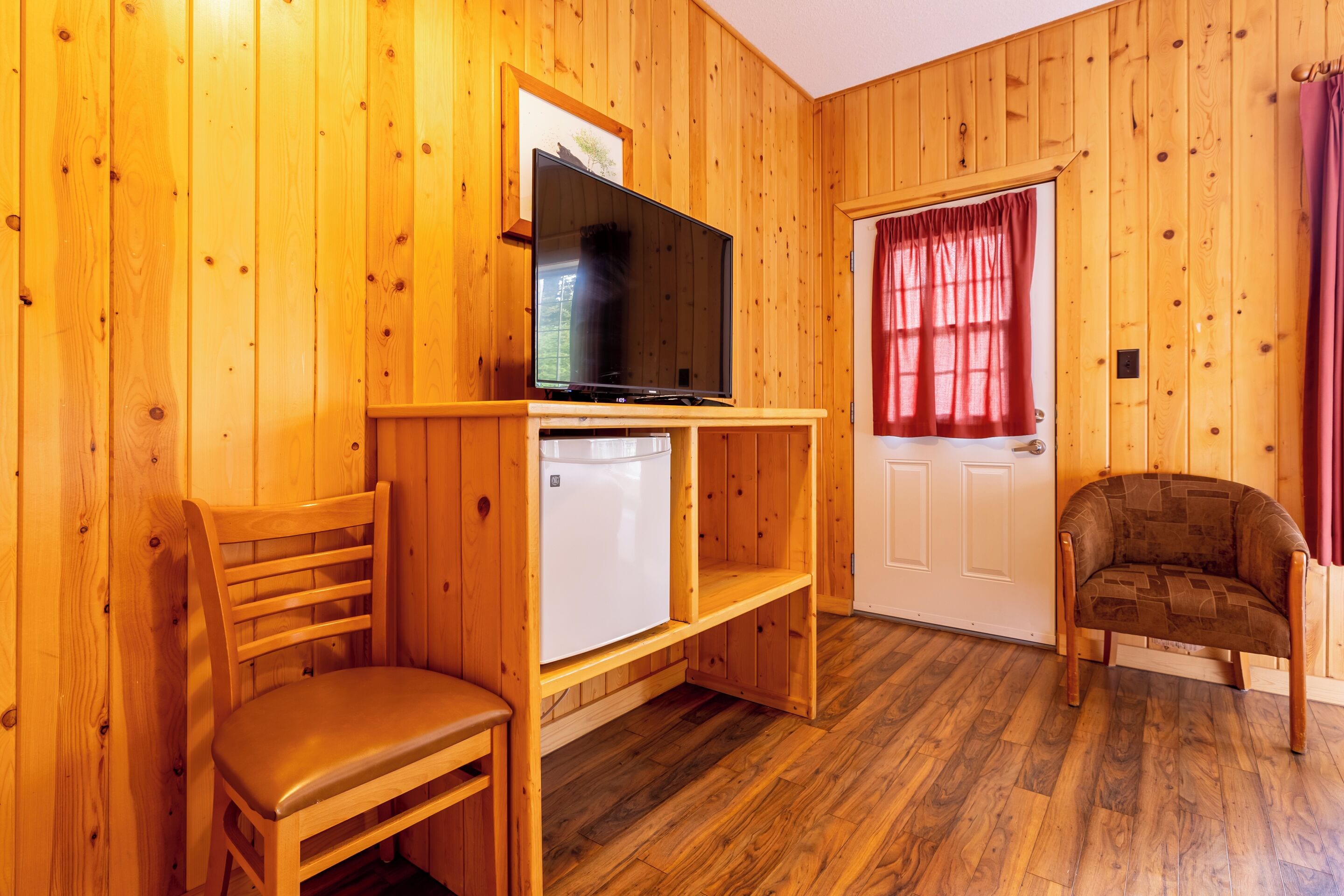 A wooden-paneled room with a television on a wooden stand, a white refrigerator, a wooden chair, and a door with a red curtain.