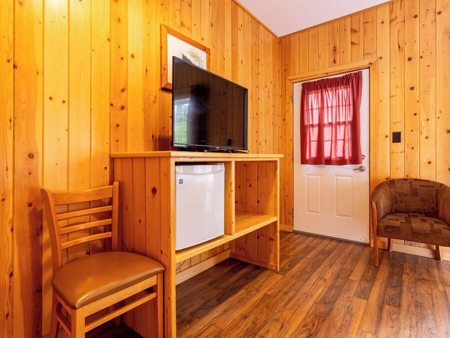 A wooden-paneled room with a television on a wooden stand, a white refrigerator, a wooden chair, and a door with a red curtain.