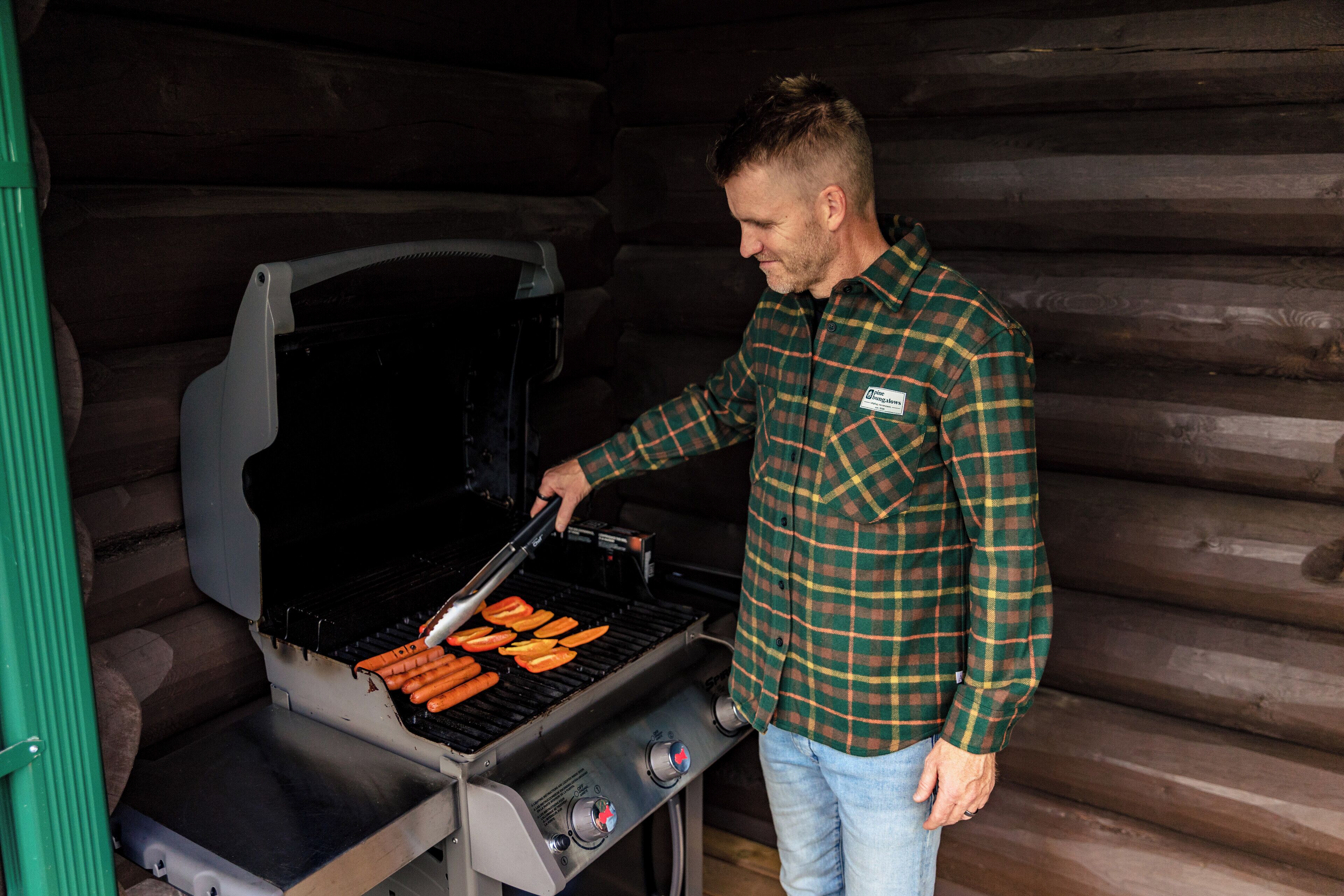 A man grilling hot dogs on an outdoor grill in a log cabin.