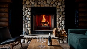 Una chimenea de piedra con fuego, una mesa de madera con copas de vino y una botella, y un sofá verde en el interior de una cabaña de troncos.