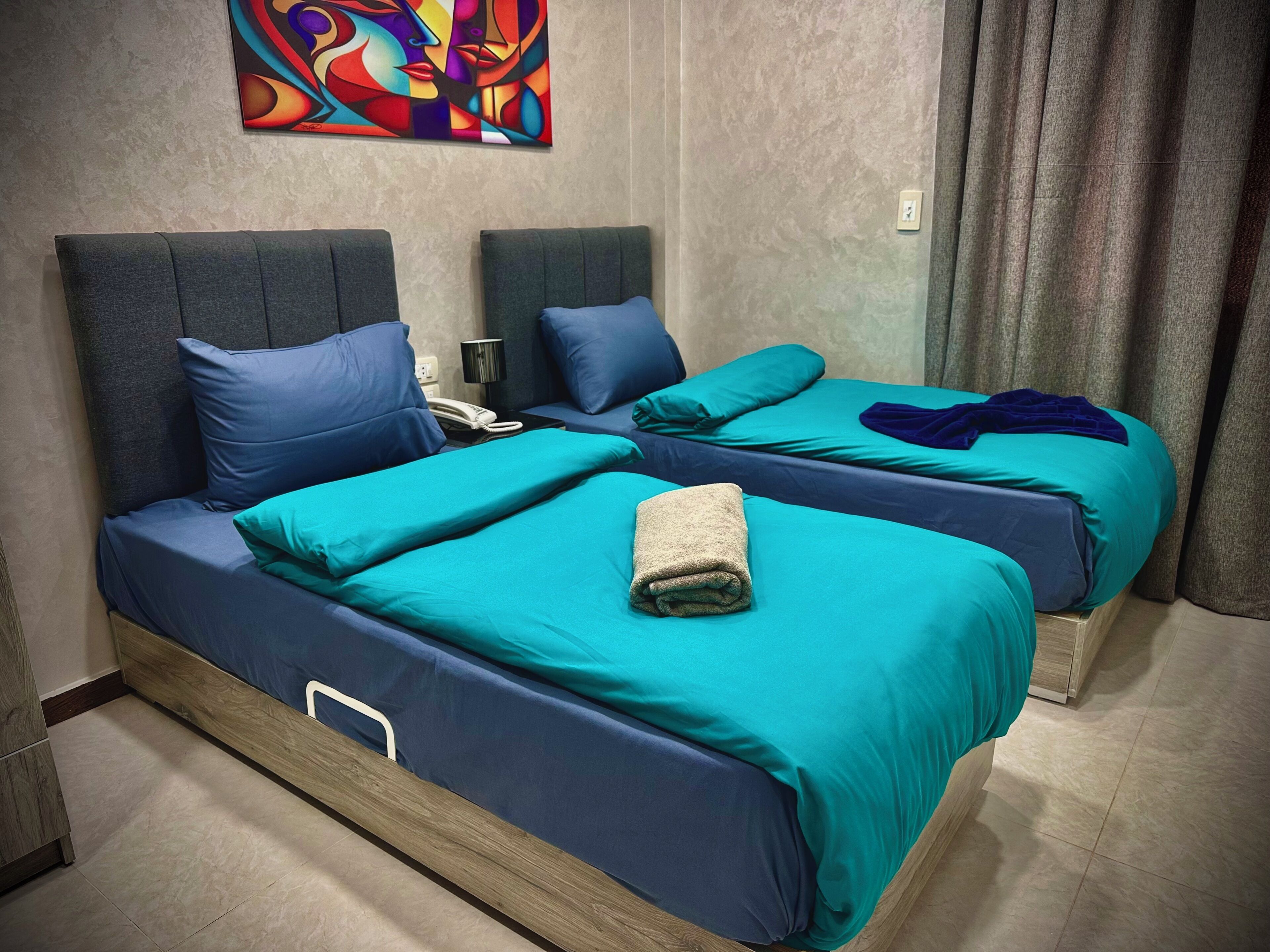 A hotel room with two beds, each with blue bedding and headboards, a wooden nightstand, a telephone, and a colorful abstract painting on the wall.