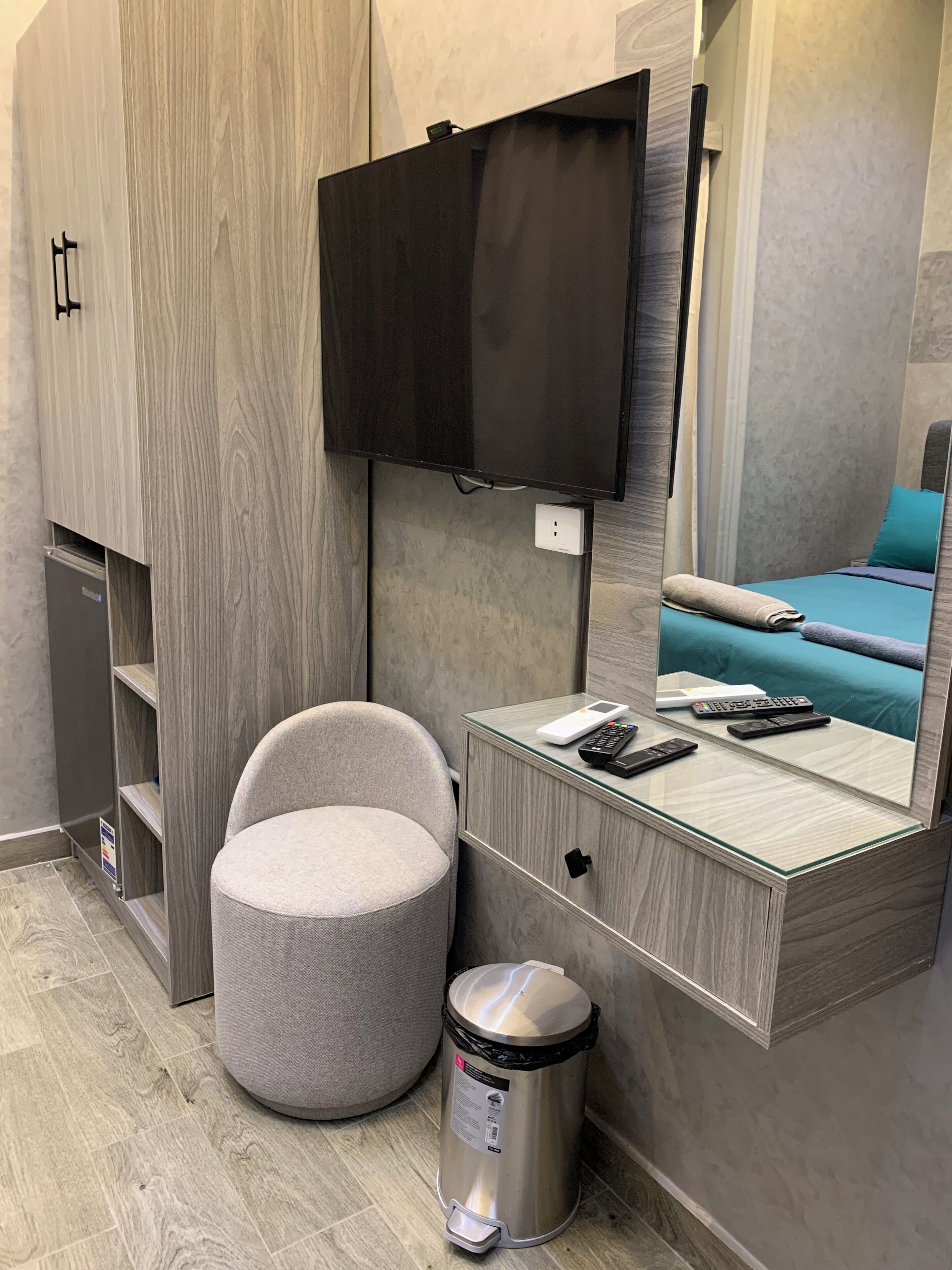 A compact hotel room with a flat-screen TV mounted on the wall, a small bedside table with a remote control, a chair, a trash bin, and a bed with a teal-colored headboard.