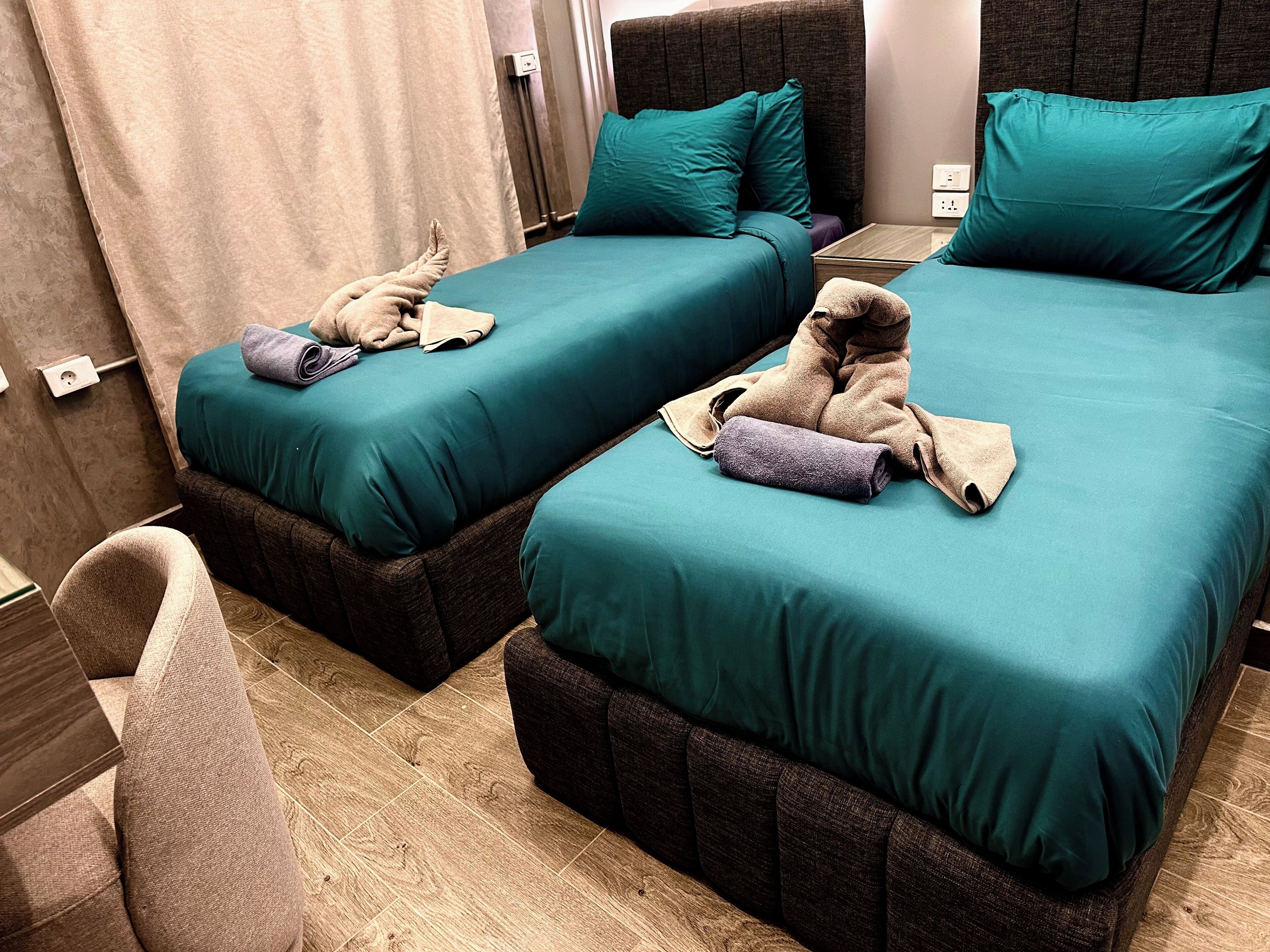 A hotel room with two beds, each with teal bedding and brown headboards. There are towels and a chair visible.