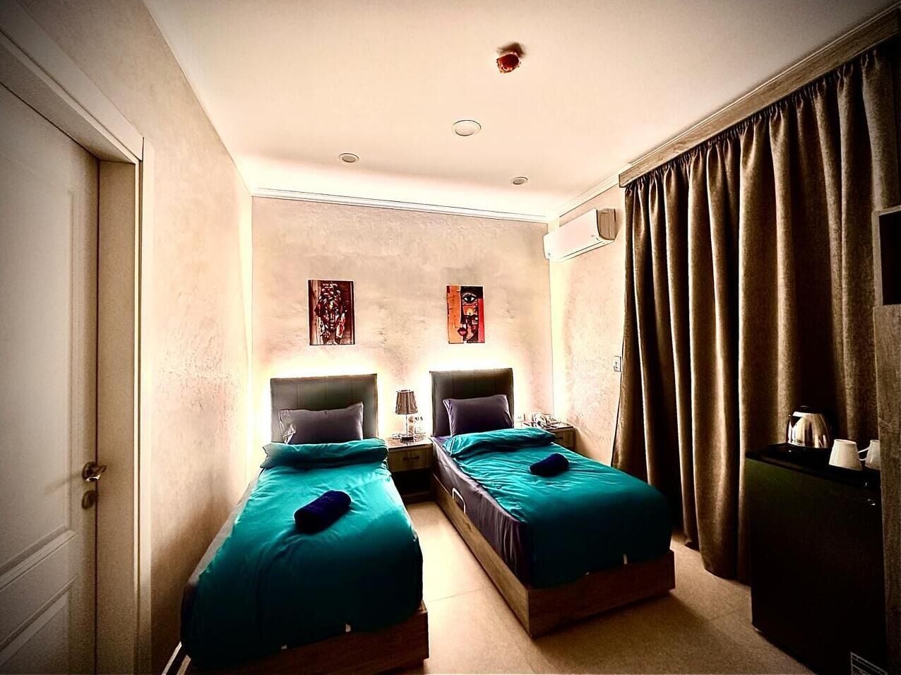 A hotel room with two beds, a nightstand with a lamp, a wall-mounted air conditioner, and a window with curtains.