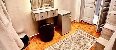 Een kleine kamer met houten vloer, een bureau met spiegel, een compacte koelkast, een prullenbak en een kamerkleed met patroon.