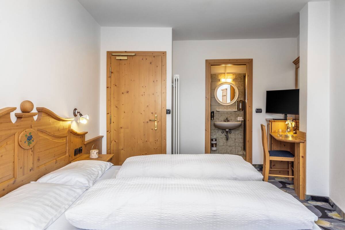 A hotel room with a wooden headboard, two beds, a wooden desk, a TV, and a bathroom with a sink and mirror.