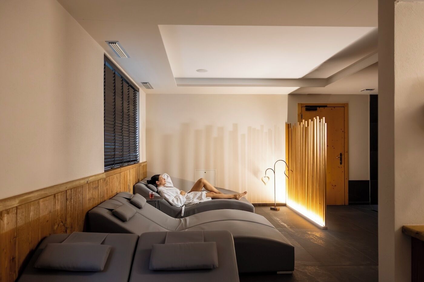 Ett spa-rum med en person som kopplar av på ett böjt massagebord. Ett massagebord syns tydligt i förgrunden, tillsammans med en trävägg, en dörr och en väggmonterad lampa.