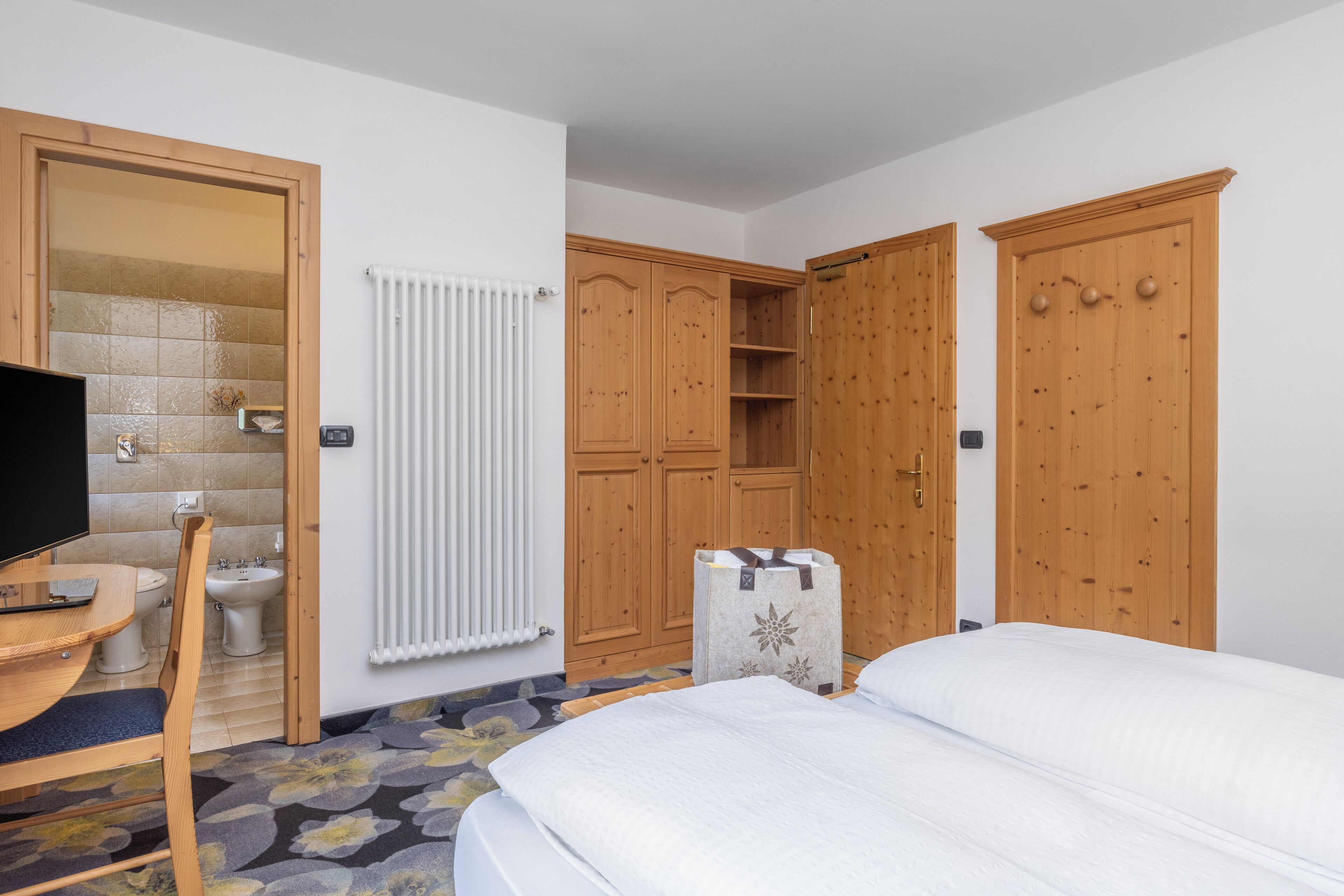 Habitación de hotel con cama, escritorio con silla, armario, radiador y televisor.