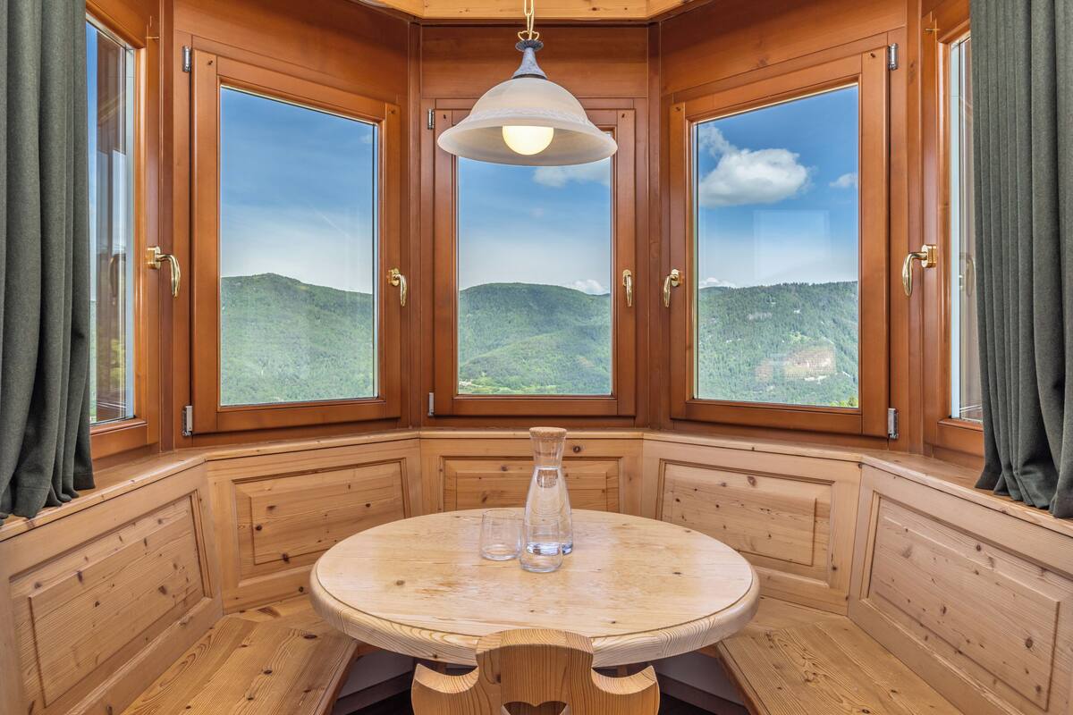 A wooden cabin with a round table and two clear glass vases, large windows with a mountain view, and a hanging light fixture.