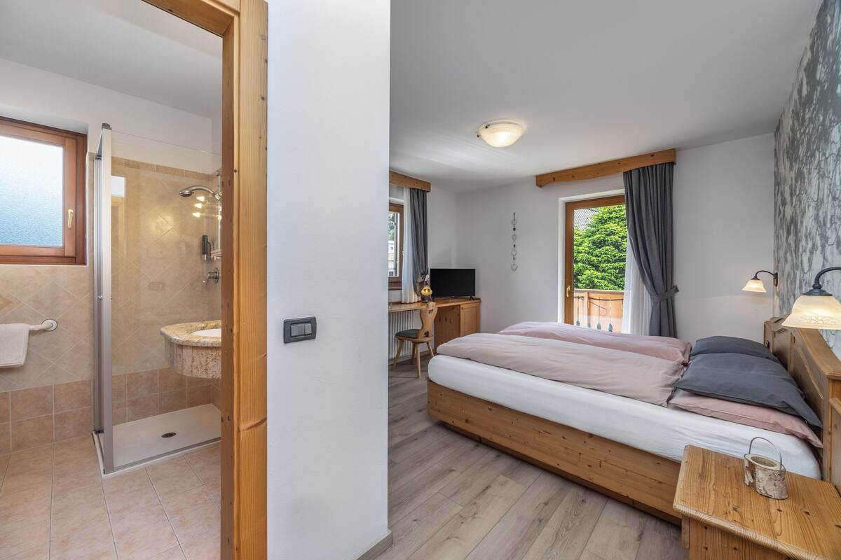 A bedroom with a wooden bed, a desk, a shower, and a view of greenery.