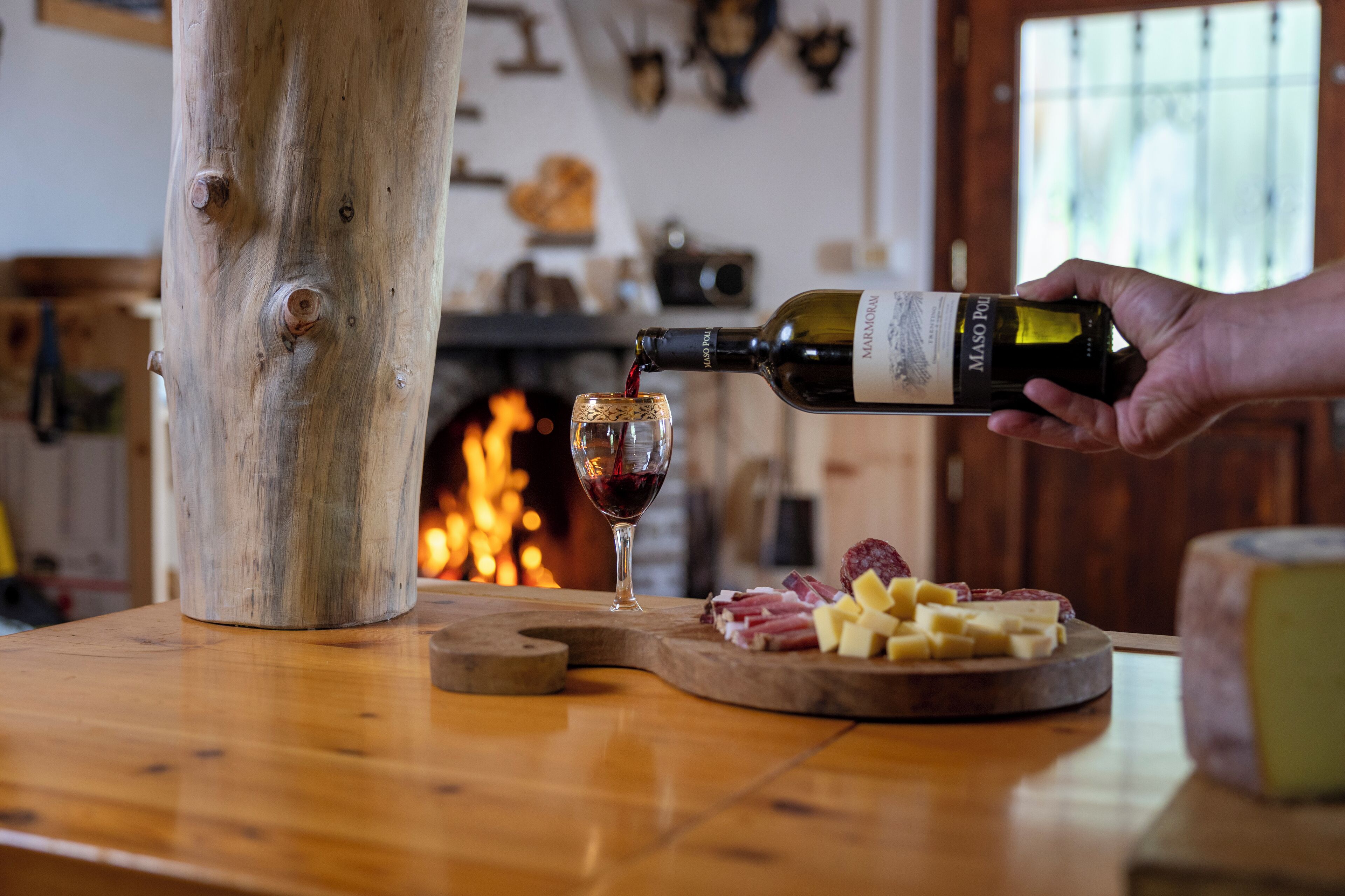 A wooden table with a bottle of wine being poured into a glass, a cheese board with various cheeses, and a fireplace in the background.