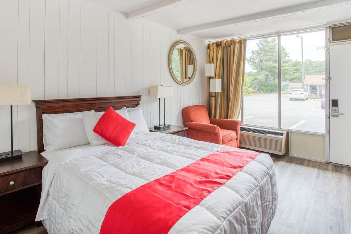 Ein Hotelzimmer mit einem großen Bett, einem Sessel, einem Fenster mit Vorhängen, einem Spiegel und einer Tür.