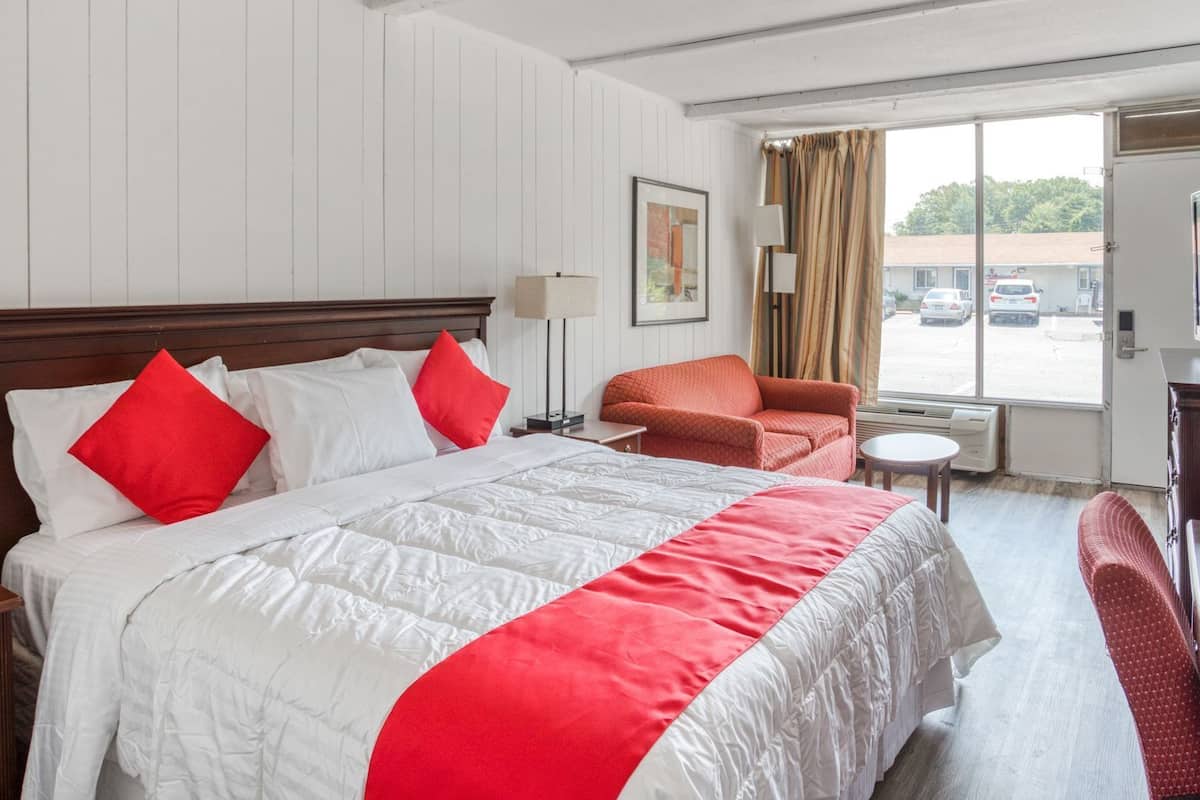 Ein Hotelzimmer mit einem großen Bett, einer rot-weißen Tagesdecke, einem roten Sessel, einem kleinen Tisch und einem Fenster mit Blick auf parkende Autos.
