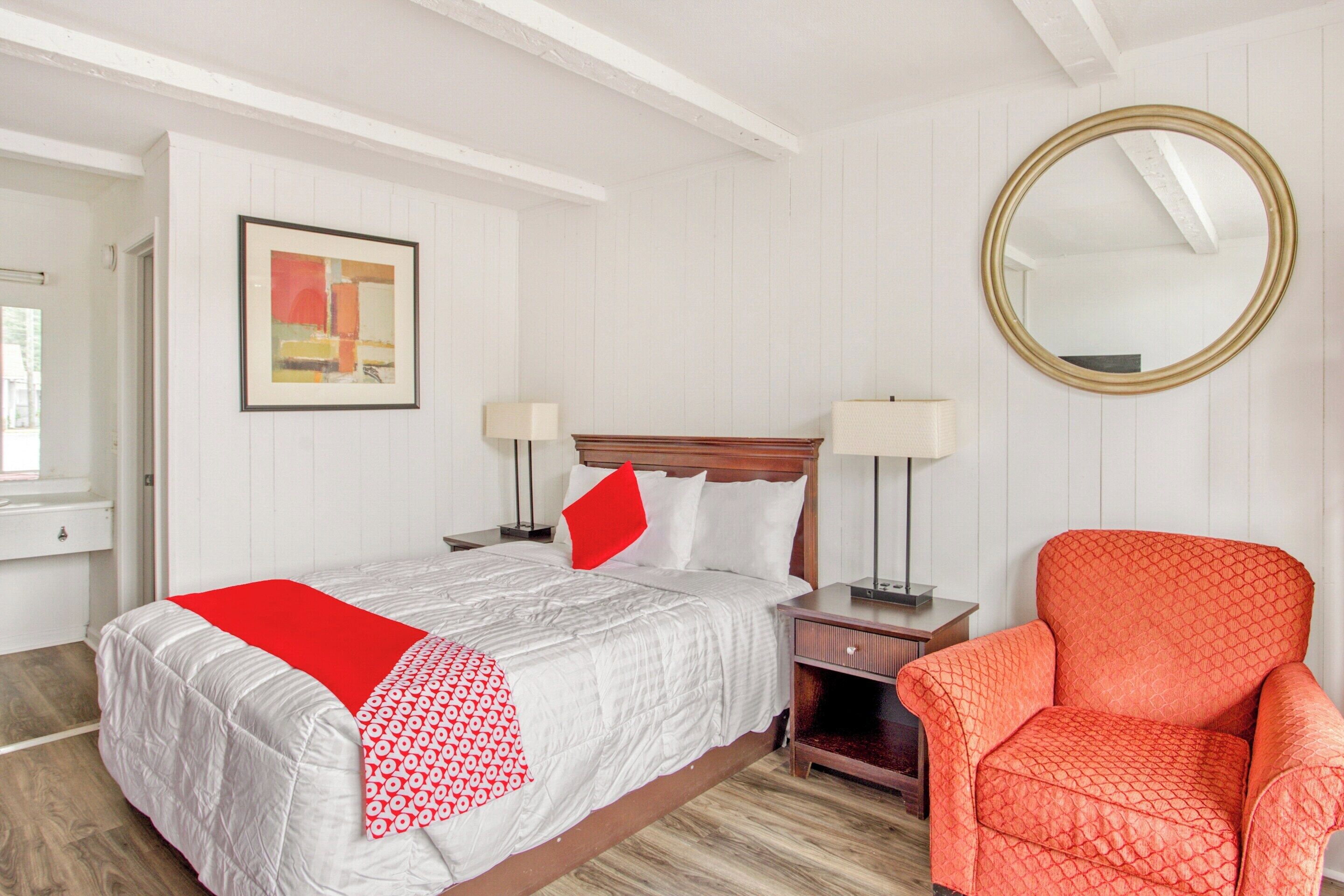 A neatly arranged hotel room with a bed, two bedside tables, a chair, a round mirror, and a framed picture on the wall.
