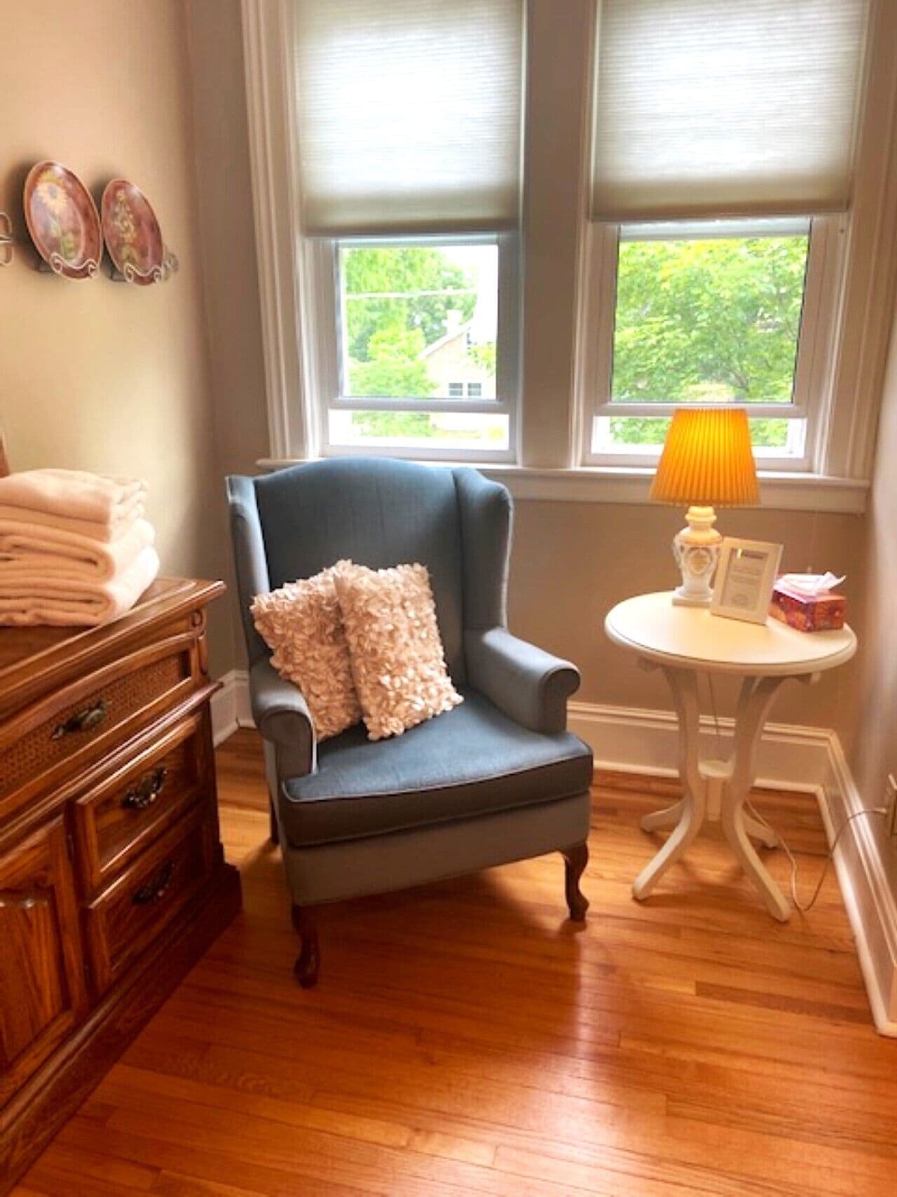 Una habitación con un sillón azul, un aparador de madera, una mesita redonda con una lámpara y una ventana con persianas.
