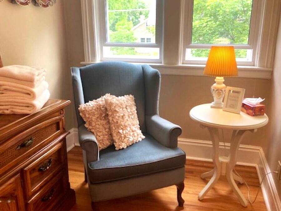 A room with a blue armchair, a wooden dresser, a small round table with a lamp, and a window with blinds.