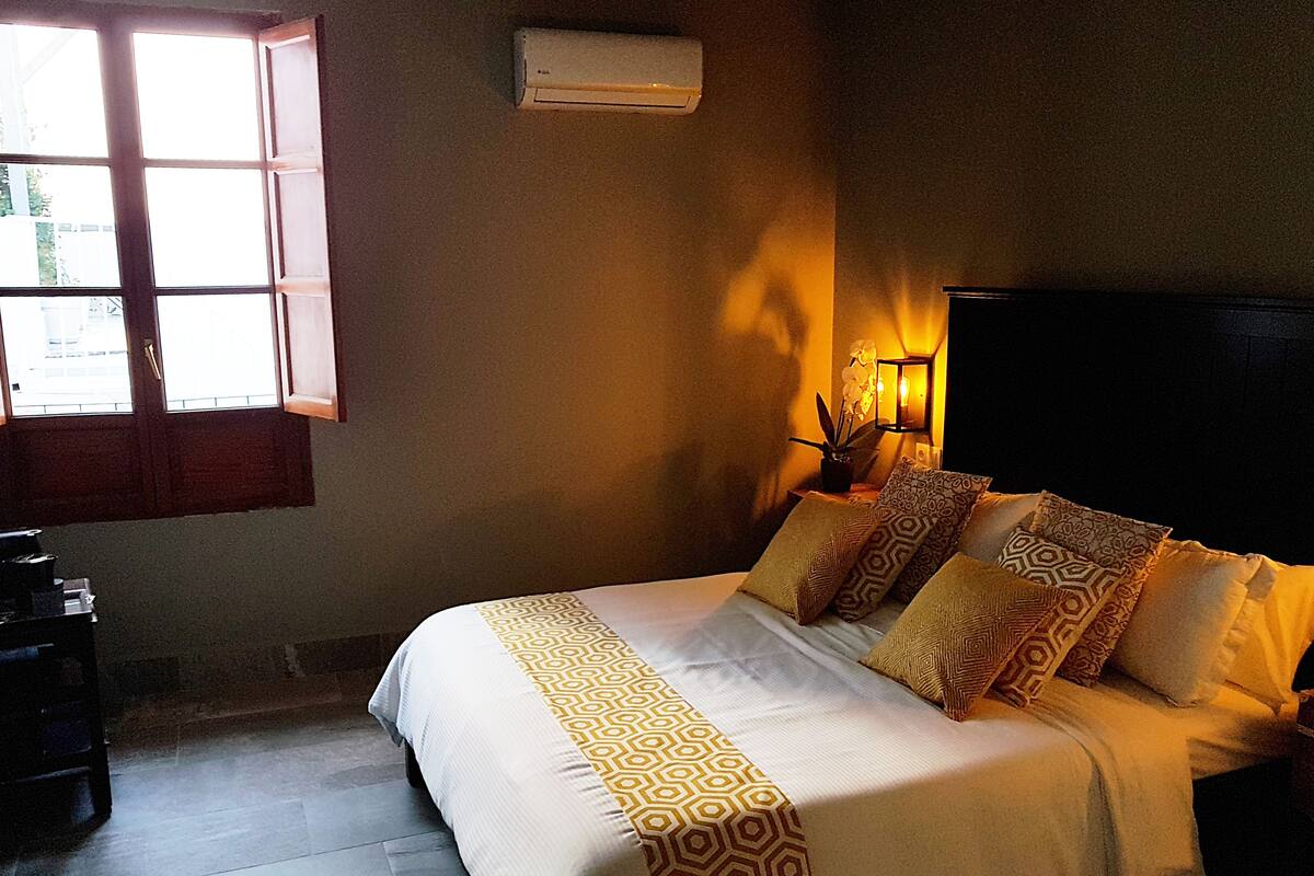 A bedroom with a bed, a window, an air conditioning unit, and a wall-mounted lamp.