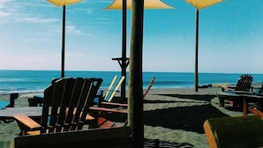 Un área de descanso frente a la playa con sillas y mesas de madera bajo sombrillas, con vista al océano.