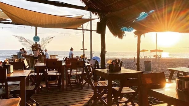 Un área de comedor al aire libre con mesas y sillas de madera, techo de paja y vista al océano al atardecer.