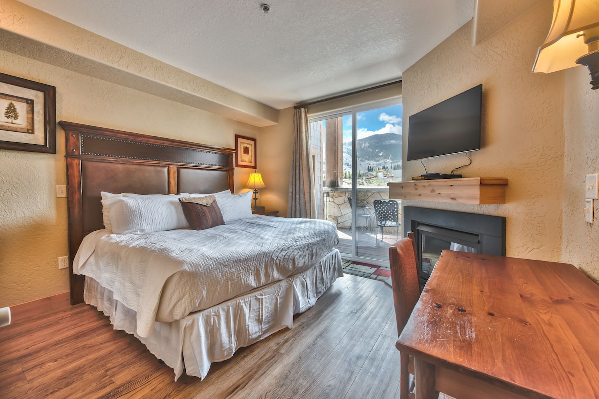 A hotel room with a large bed, a wooden table, a fireplace, and a TV mounted on the wall.