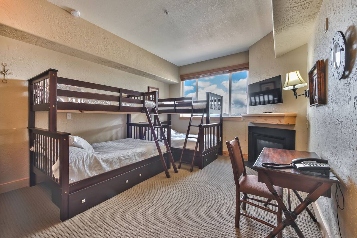 A room with bunk beds, a desk with a chair, a fireplace, and a TV mounted on the wall.