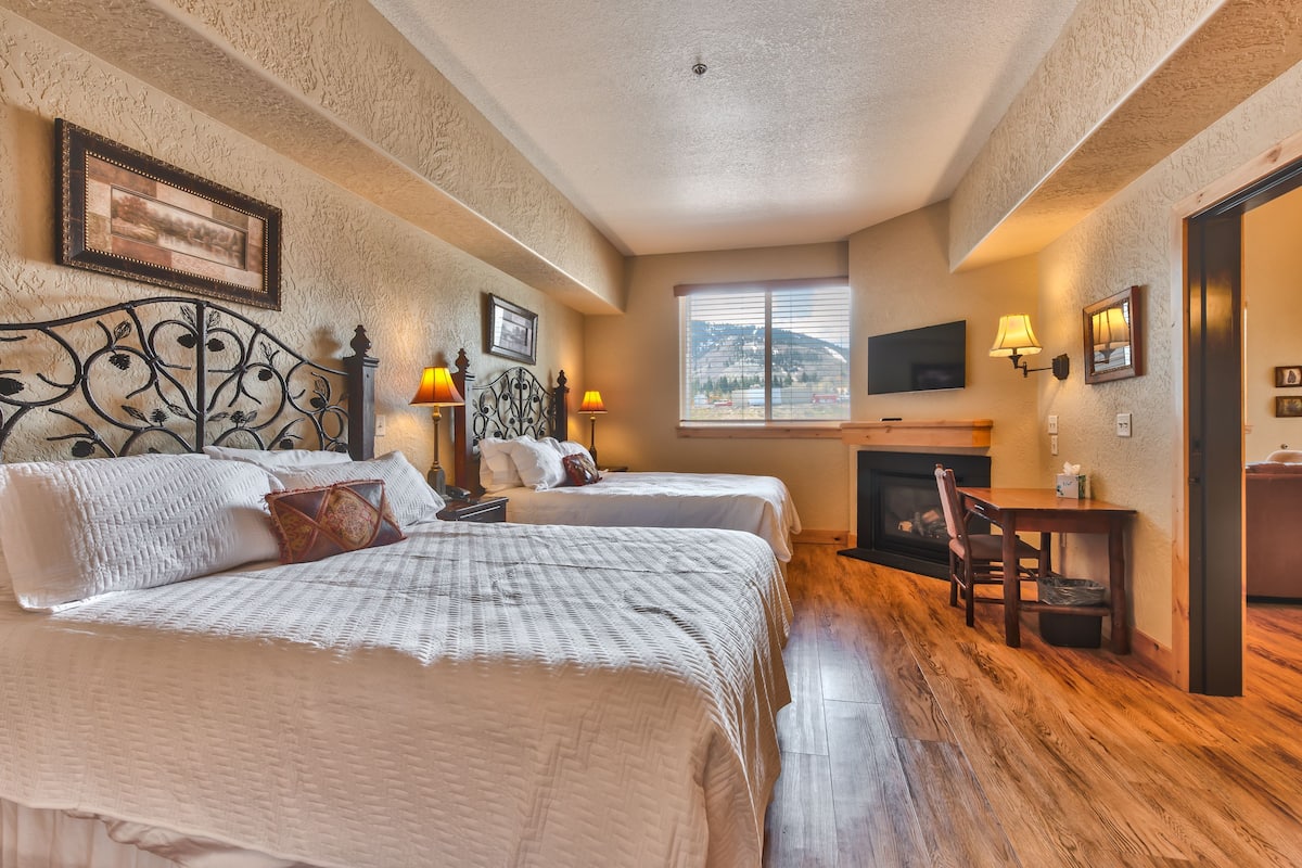 A hotel room with two beds, a fireplace, a TV, and a window with a mountain view.