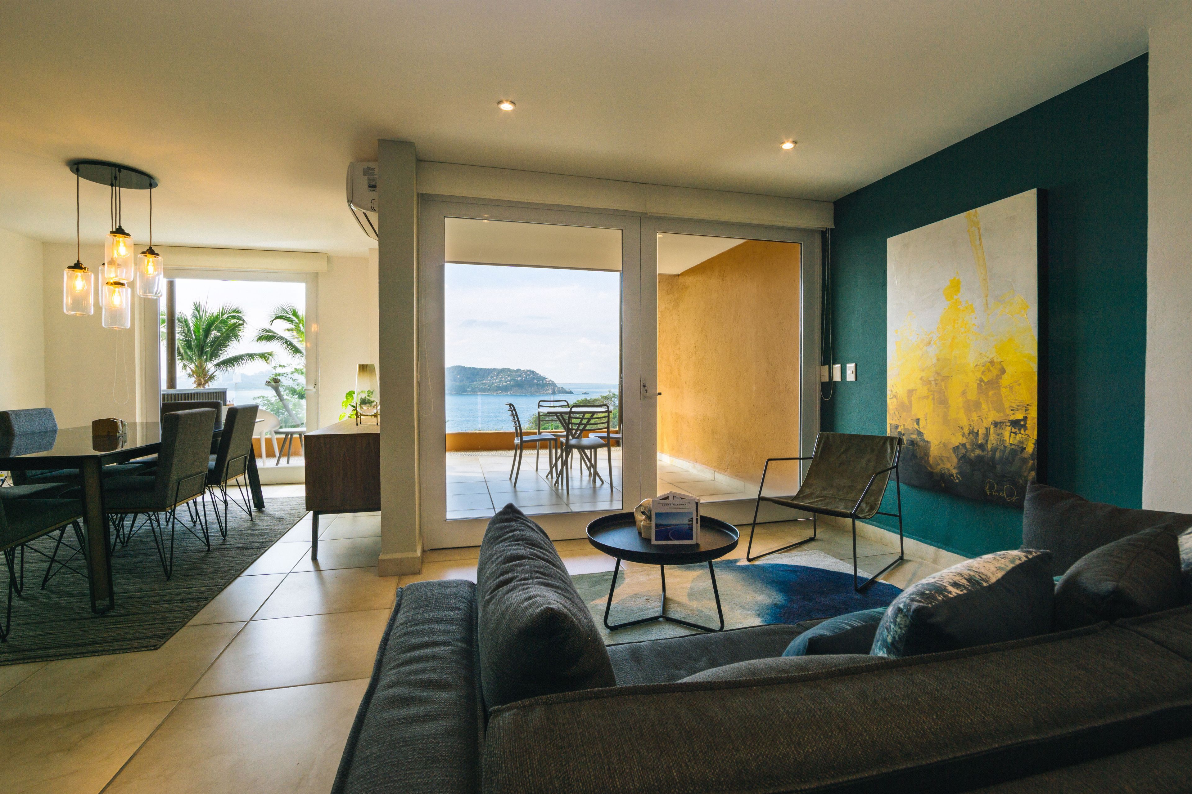 A modern living room with a sofa, dining table, chairs, and a view of the sea through large glass doors.