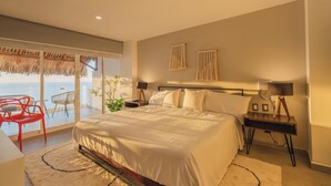 Ein Schlafzimmer mit einem großen Bett, einem Nachttisch mit Lampe, Blick auf das Meer durch eine Glastür und einem roten Sessel.